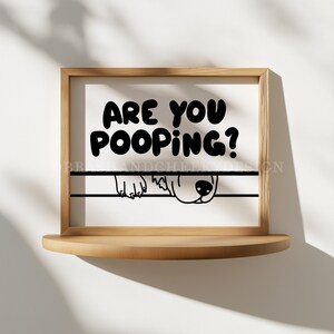 May include: A framed print with the text "ARE YOU POOPING?" in bold black letters. Below the text, a black line and a cartoon dog's face and paws peek out. The frame is light brown, and the print sits on a wooden shelf.