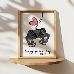 Könnte beinhalten: Ein gerahmter Druck mit einem Holzrahmen, der ein rosa Herz, einen Videospiel-Controller und zwei graue Fußabdrücke zeigt. Der Text lautet "we you" und "happy father's day 2025". Der Druck wird auf einem Holzregal angezeigt.