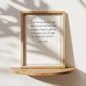 May include: A framed print with a light wood frame and a white background. The print features a quote from John 14:27 in black text. The frame rests on a small, curved wooden shelf.