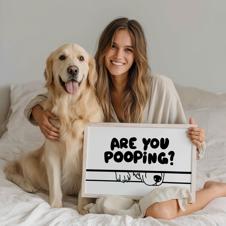 May include: A smiling woman and a golden retriever dog are seated on a bed. The woman holds a framed sign with the text "ARE YOU POOPING?" in black letters. The dog's head and paws are illustrated below the text.