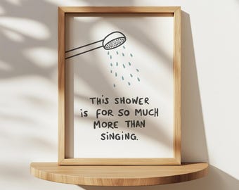 May include: A framed print with a light wood frame, featuring a showerhead illustration and the text: "THIS SHOWER IS FOR SO MUCH MORE THAN SINGING." The print is displayed on a wooden shelf against a white wall.