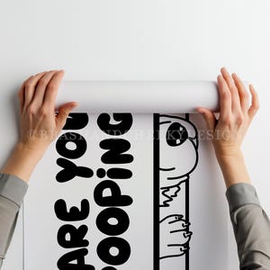 May include: A white poster with black text and a cartoon alien illustration is being unrolled. The text reads "Are You Kidding?" The alien is peeking out from behind a vertical line. The poster is held by two hands.