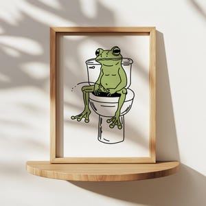 Può includere: Una stampa artistica incorniciata con un cartone animato di una rana verde seduta su un water. La rana ha un'espressione rilassata e l'opera è delineata in nero. La cornice è in legno chiaro e lo sfondo è bianco.