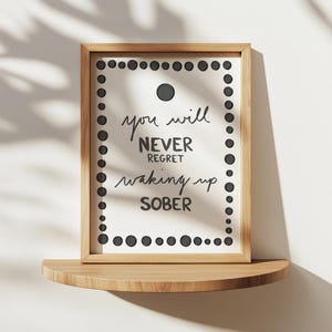 May include: A framed print with the text "you will NEVER regret waking up SOBER" in black script. The frame is light brown, and the print has a border of black dots. The print is displayed on a wooden shelf.