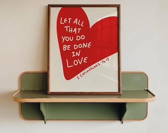 May include: A framed print featuring a red heart and the white text "LET ALL THAT YOU DO BE DONE IN LOVE". The print is displayed on a two-tiered green and beige shelf, mounted on a white wall. The text "1 Corinthians 16:14" is also visible.
