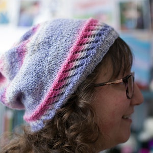 May include: A woman wearing a purple and pink knitted hat with a lacy design. The hat is slouched on her head and she is looking to the side.