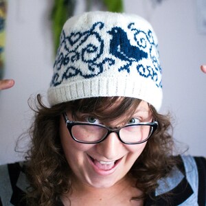 May include: A woman wearing a white knitted hat with a blue bird design and black framed glasses. She is smiling and pointing at her hat.