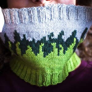 May include: A knitted cowl in shades of gray, green, and chartreuse. The cowl features a forest-like pattern in dark green and chartreuse against a gray background. The bottom edge is a bright chartreuse color.