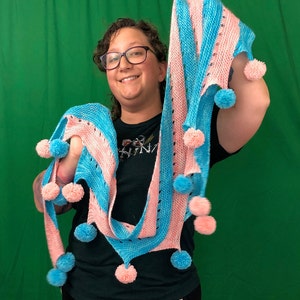 May include: A person wearing a blue and pink striped scarf with pom-poms. The scarf is crocheted and has a light blue and pink color scheme.