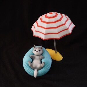 May include: A small gray and white husky dog figurine is laying in a blue bean bag chair with a red and white striped beach umbrella behind it.