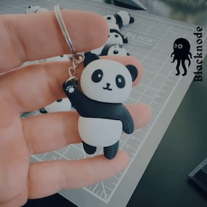 Puede incluir: Un llavero de panda blanco y negro con un anillo de llave plateado. El panda está levantando su pata y tiene una expresión linda en su rostro.