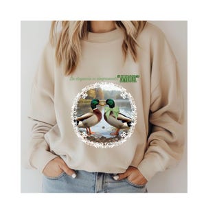 May include: Beige sweatshirt featuring a circular graphic of two ducks with green bows, kissing. The graphic is surrounded by a white floral design. Above the graphic is the text "La elegancia es simpremente "AMOR".