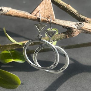 May include: Silver hoop earrings with a triangle charm. The hoops have a textured design and are connected to the triangle charm by a small loop.