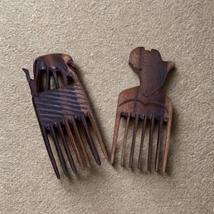 May include: Two wooden hair combs with detailed carvings. One comb has an elaborate design, the other a stylized human figure. Both are made of dark wood and have multiple teeth, showcasing craftsmanship.