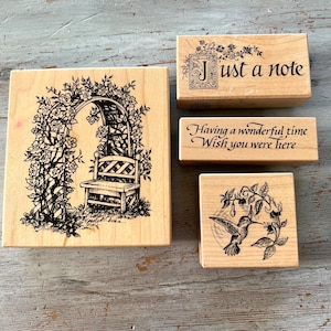 May include: Four wooden rubber stamps with floral designs. The first stamp features a rose trellis with a bench. The second stamp says "Just a note" with a decorative letter J. The third stamp says "Having a wonderful time Wish you were here". The fourth stamp features a hummingbird with flowers.