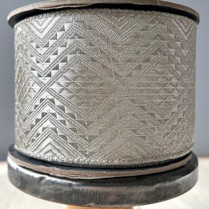 Op de afbeelding: Een spoel zilveren lint met een geometrisch patroon. Het lint is gewikkeld om een zwarte metalen spoel en staat op een houten basis.