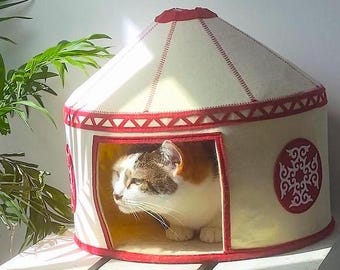 Puede incluir: Un gato está dentro de una cama para mascotas de estilo yurt de color crema con adornos y detalles decorativos rojos. La cama para gatos tiene un diseño circular con una parte superior roja y una abertura rectangular. El gato es blanco, marrón y naranja.