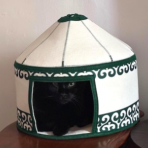 May include: A circular pet bed shaped like a yurt with a cream-colored roof and walls. The structure features dark green trim and a repeating dark green scroll pattern along the base. A black cat is visible inside the square front opening.
