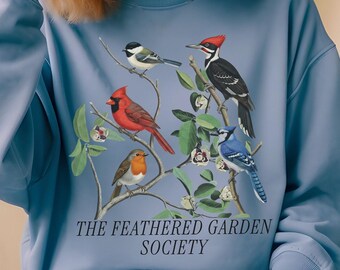 May include: A dusty blue crewneck sweatshirt featuring a graphic print of various birds perched on branches, including a cardinal, robin, and woodpecker. The text "THE FEATHERED GARDEN SOCIETY" is printed in a dark serif font below the bird illustration.