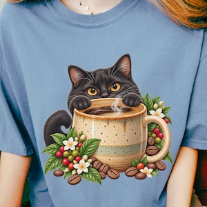 Op de afbeelding: Een blauw t-shirt met korte mouwen en een afbeelding van een zwarte kat die over de rand van een gespikkelde beige koffiemok kijkt. De mok is omringd door groene koffiebladeren, witte bloesems, rode koffiebessen en bruine koffiebonen.