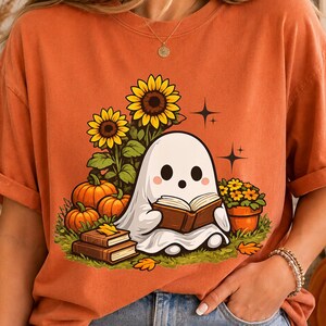 May include: An orange short-sleeve t-shirt featuring a graphic print of a white ghost reading a book. The illustration includes yellow sunflowers, orange pumpkins, a potted plant, and stacked books on a green grassy patch with small star accents.