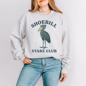 May include: Light gray sweatshirt with the text "SHOEBILL STARE CLUB" above and below a graphic of a shoebill stork. The bird is dark gray with a large yellow beak and stands on two thin legs.