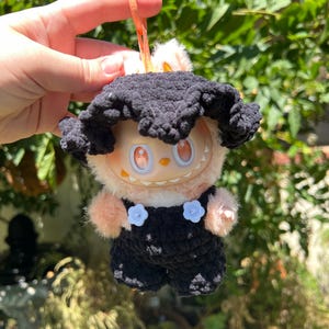 Op de afbeelding: Een pluche pop met een zwarte gehaakte hoed en tuinbroek, met een perzikkleurig gezicht en armen. De tuinbroek heeft witte bloemvormige knopen. De pop wordt vastgehouden door een oranje sleutelhanger.