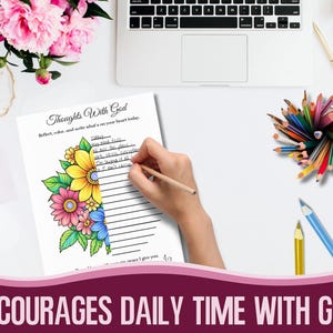 May include: A journal page titled "Thoughts With God" with floral illustrations. A hand holding a pencil is writing on the page. The page is surrounded by coloured pencils, a laptop, and pink flowers.
