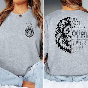 May include: A light gray crew neck t-shirt featuring a black lion graphic and text. The front of the shirt displays a lion's head and the words "LION OF JUDAH". The back of the shirt has a lion's head and the text "DO NOT WEEP. THE LION OF THE TRIBE OF JUDAH".