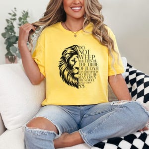 May include: Yellow t-shirt featuring a black lion graphic with text. The text reads "Do Not Weep, The Lion of the Tribe of Judah Has Triumhed So That He May Open The Scroll Revelation 5:5". The shirt is worn by a person.