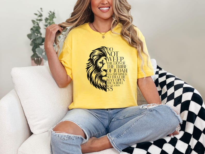 May include: Yellow t-shirt featuring a black lion graphic with text. The text reads "Do Not Weep, The Lion of the Tribe of Judah Has Triumhed So That He May Open The Scroll Revelation 5:5". The shirt is worn by a person.