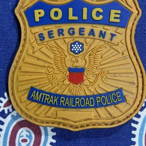 May include: A gold-colored police badge with a blue background. The badge features an eagle with a shield and the text "POLICE SERGEANT AMTRAK RAILROAD POLICE".