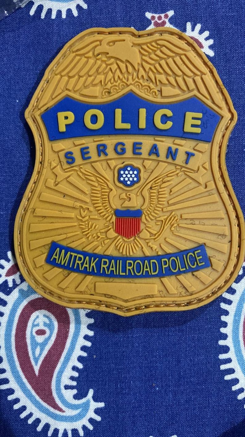 May include: A gold-colored police badge with a blue background. The badge features an eagle with a shield and the text "POLICE SERGEANT AMTRAK RAILROAD POLICE".