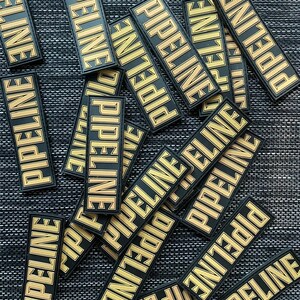 May include: A pile of black and gold patches with the word "PIPELINE" printed on them. The patches are arranged in a random pattern on a dark gray textured surface.