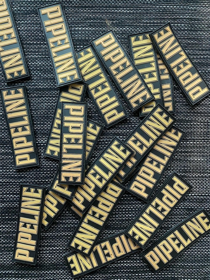May include: A pile of black and gold patches with the word "PIPELINE" printed on them. The patches are arranged in a random pattern on a dark gray textured surface.