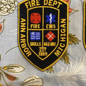 May include: A black and gold embroidered patch with the text "FIRE DEPT ANN ARBOR MICHIGAN" and the text "EST 1869". The patch has four smaller squares with the text "FIRE", "EMS", "SKILLS", and "HAZ-MAT".
