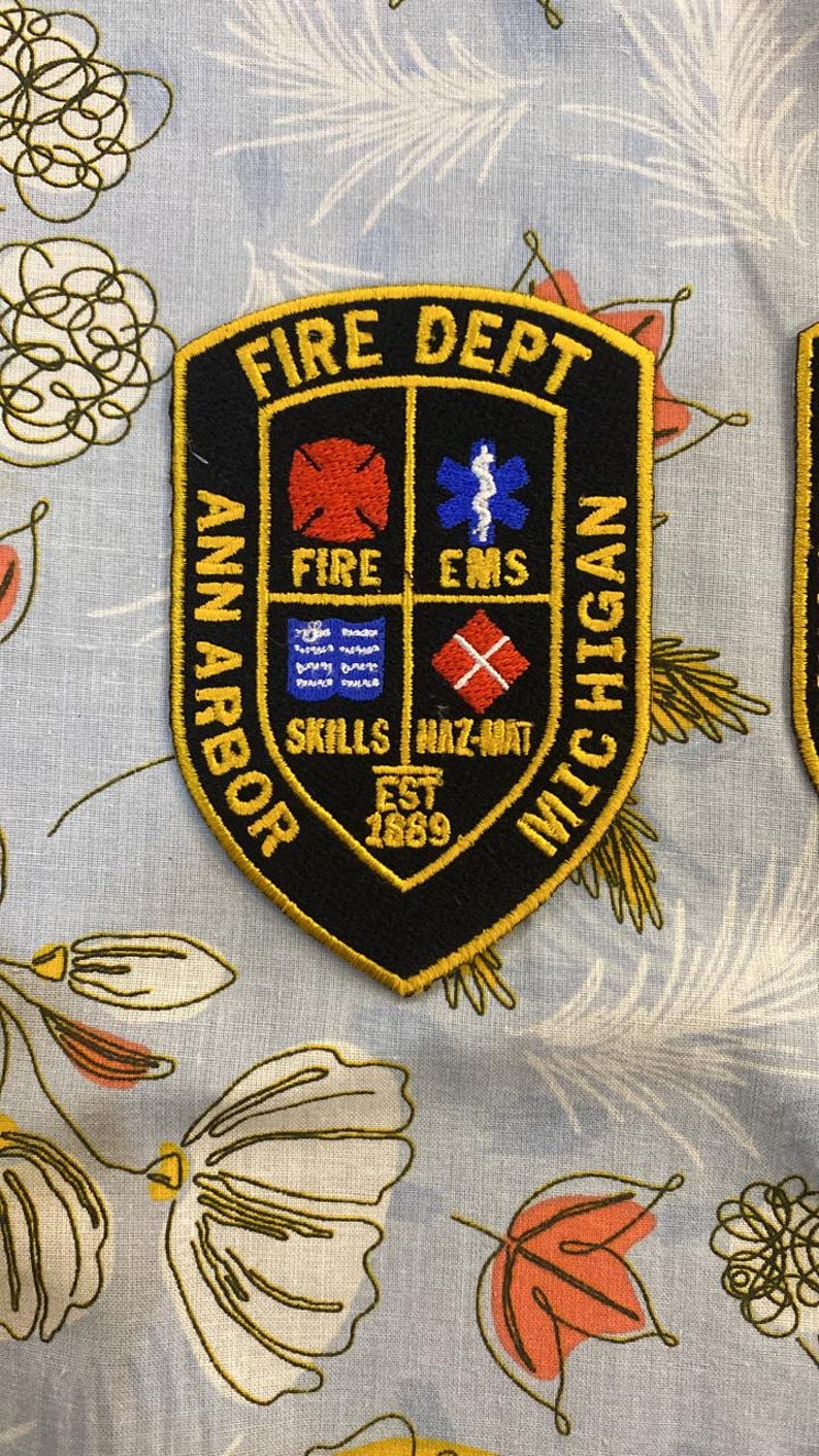 May include: A black and gold embroidered patch with the text "FIRE DEPT ANN ARBOR MICHIGAN" and the text "EST 1869". The patch has four smaller squares with the text "FIRE", "EMS", "SKILLS", and "HAZ-MAT".