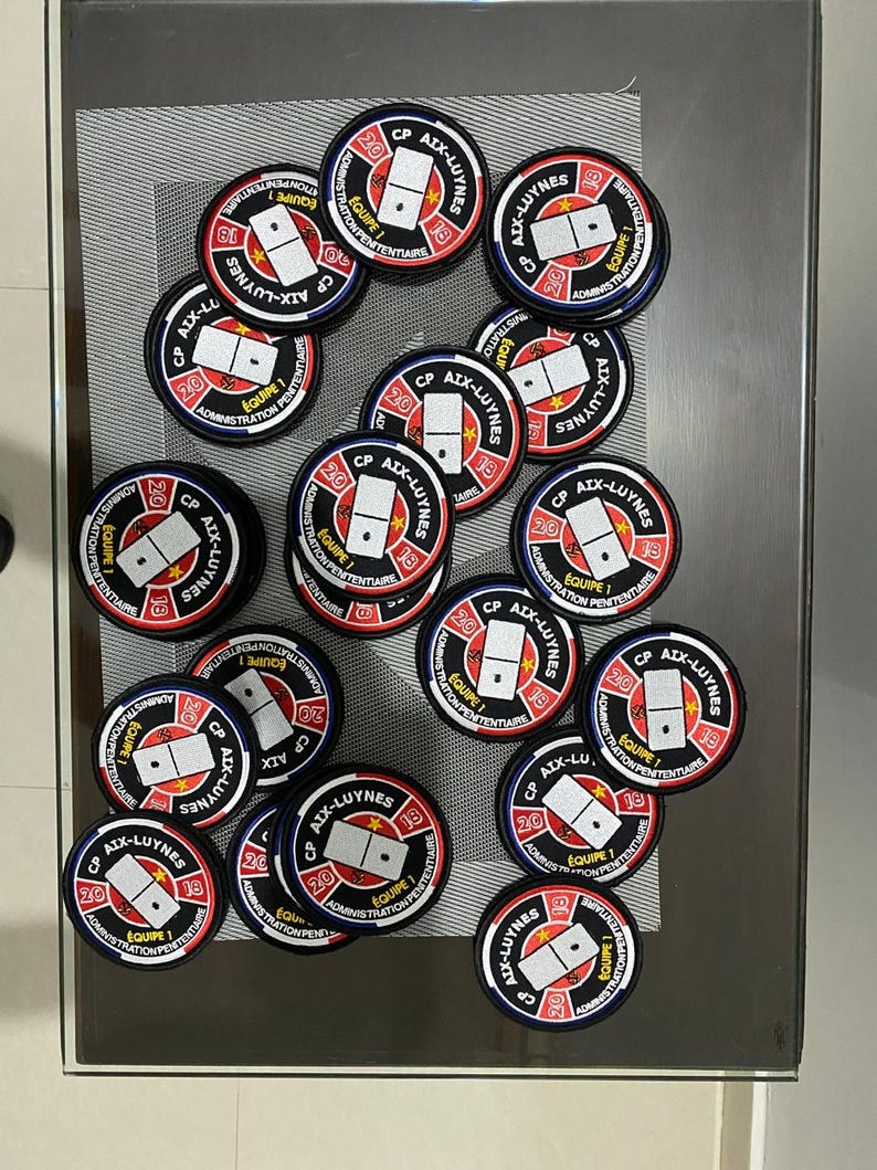 May include: A collection of 18 round black and white patches with red accents. Each patch features a domino with the text "CP AIX-LUYNES" and "&Eacute;QUIPE 1" in French. The patches are arranged in a circular pattern on a glass table.