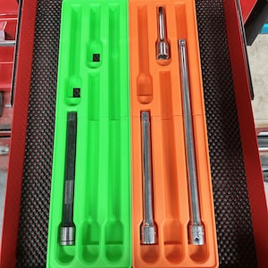 Peut inclure: Tiroir de boîte à outils rouge avec deux plateaux en plastique contenant divers outils métalliques. Un plateau est vert, l'autre orange. Les outils comprennent des douilles et des rallonges. Le tiroir a une doublure noire texturée.