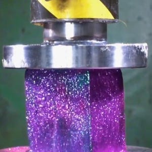 May include: A close-up of a purple, metallic, hexagonal object being pressed by a metal press. The object is being compressed from the top and bottom.
