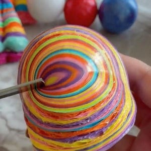 May include: A colorful, rainbow-striped, sphere-shaped object made of layered paper. The object is being held in a hand and a small tool is being used to puncture the surface.