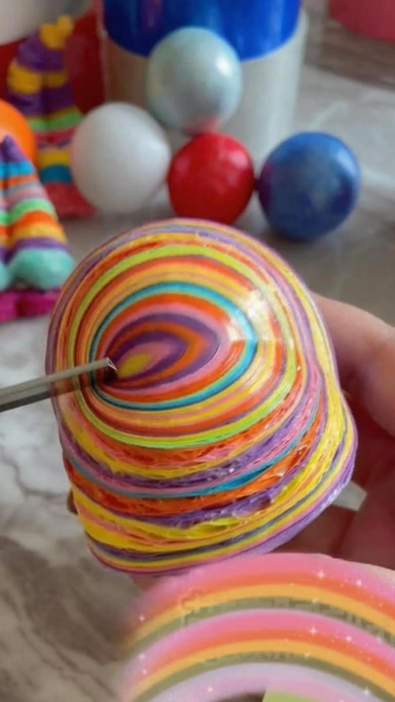 May include: A colorful, rainbow-striped, sphere-shaped object made of layered paper. The object is being held in a hand and a small tool is being used to puncture the surface.