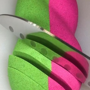 May include: A green and pink kinetic sand ball being sliced with a knife. The sand is cut into thin slices.
