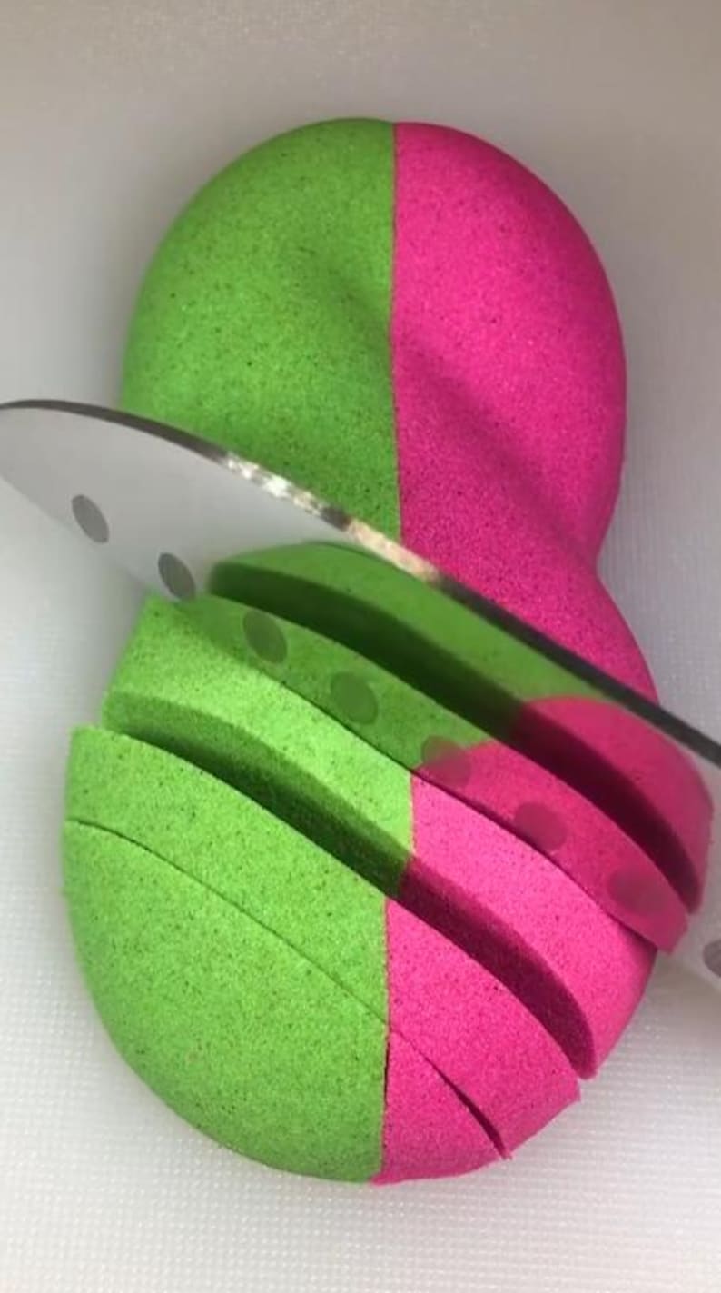 May include: A green and pink kinetic sand ball being sliced with a knife. The sand is cut into thin slices.
