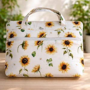 May include: White laptop bag featuring a sunflower design. The bag has a zip closure and a handle. The sunflowers are yellow and brown with green leaves, set against a white backdrop. The bag is on a wooden surface.