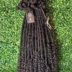 May include: Black dreadlocks hair extensions with curly ends. The hair is displayed against a green background. The label reads "Pearl Custom Locks".