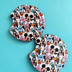 May include: Two circular car cup holder coasters featuring a repeating pattern of cartoon character faces including Mickey Mouse, Minnie Mouse, Donald Duck, Daisy Duck, and Goofy on a white background, each with a small semi-circular notch on the side.