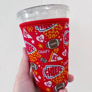 May include: A red fabric cup sleeve featuring a repeating pattern of footballs, white pennants with KC text, hearts, yellow leopard spots, and the word Chiefs in white and red lettering. The sleeve has a red stitched trim at the top edge.
