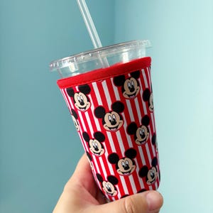 Puede incluir: Un vaso de plástico transparente con pajita, que incluye una funda de tela decorada con un patrón repetitivo de caras de Mickey Mouse sobre rayas verticales rojas y blancas. La funda tiene un borde de tela roja en la parte superior.