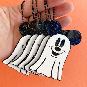 May include: A collection of Halloween-themed keychains featuring a smiling ghost design with Mickey Mouse ears. The ghosts are white with black outlines, and the ears are dark blue with white speckles. Each keychain has a black ball chain.